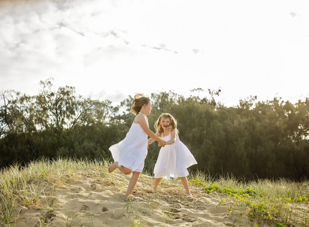 2 girls dancing on top of a sand dune on the beach smiling