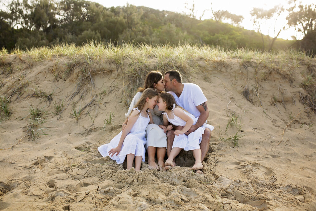 a family of four sitting on the beach, mum and dad are kissing