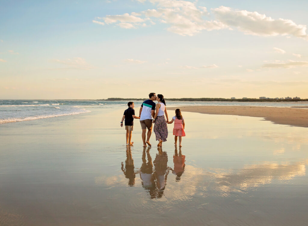 family of 4 walking along the beach with parents kissing