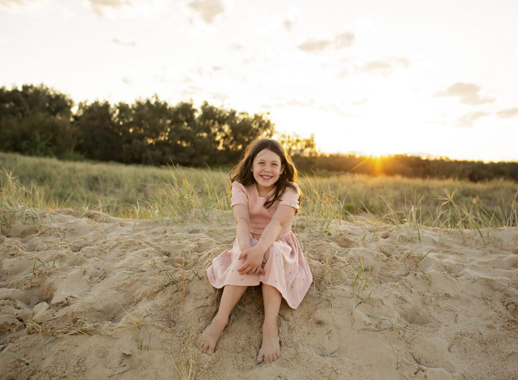 girl sitting on a sand dune smiling with the sunset behind her