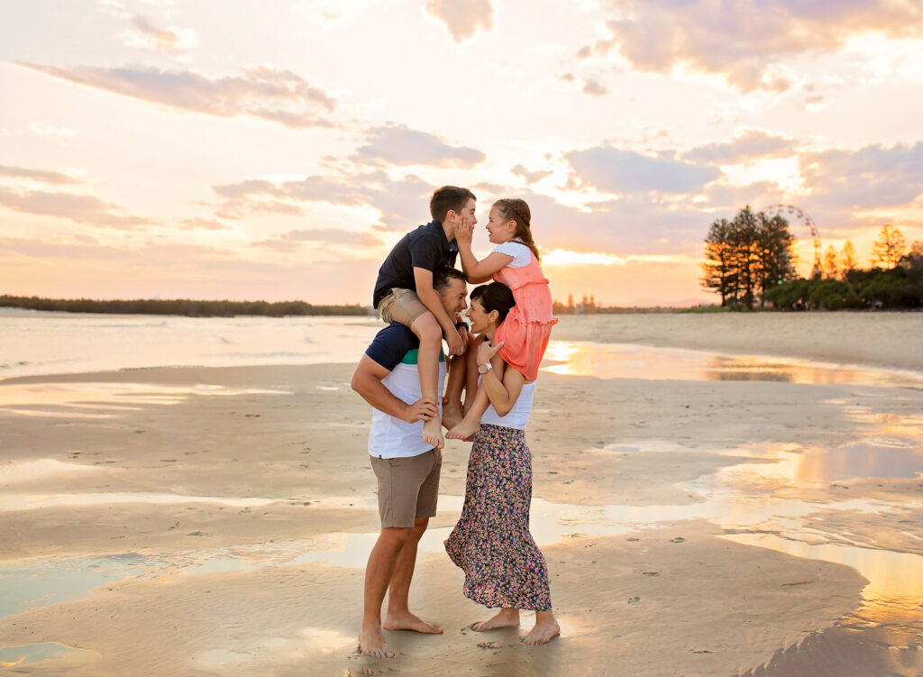 mum and dad with kids on their shoulders laughing on the beach