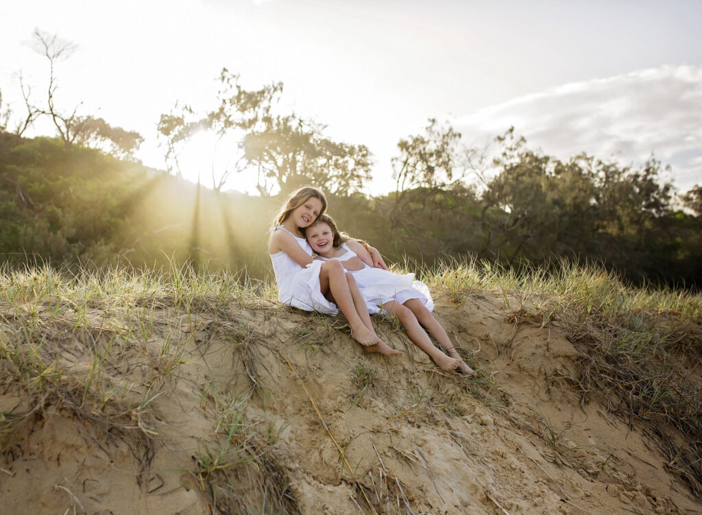 2 sisters sitting on a sand dune with the sunset and rays behind them