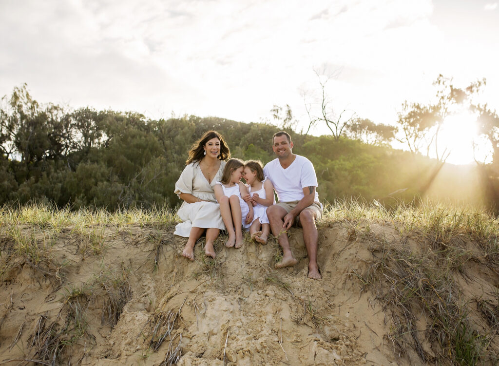 family of four sitting on a sand dune with the sunset and rays behind them