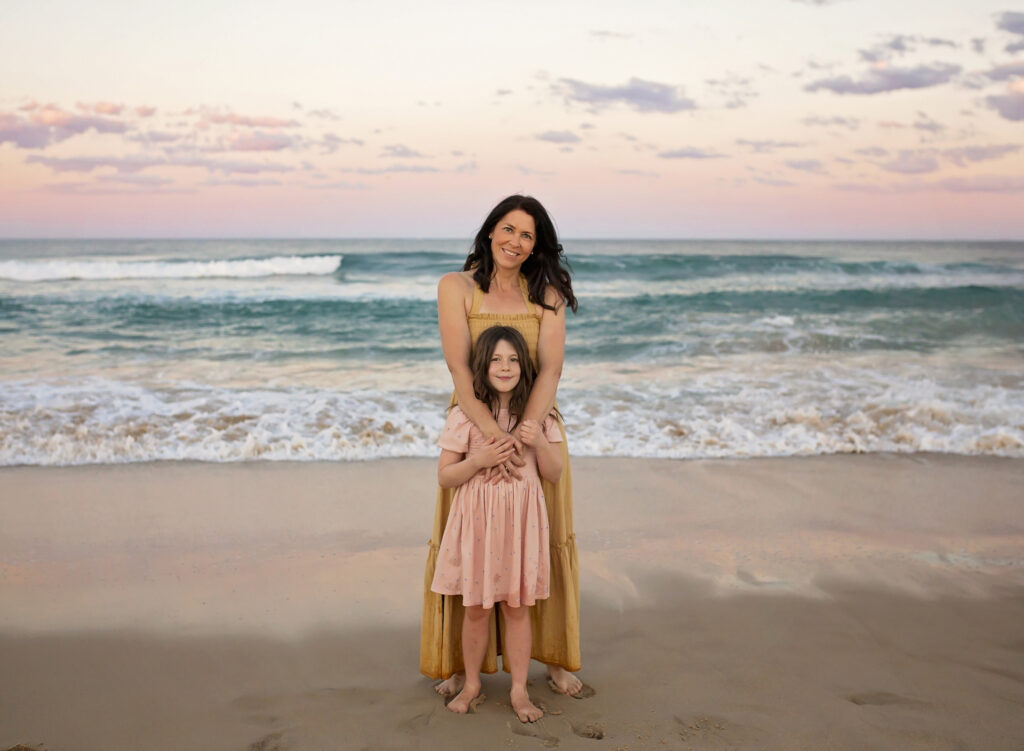 a mother and her daughter standing on the sand with the ocean behind them