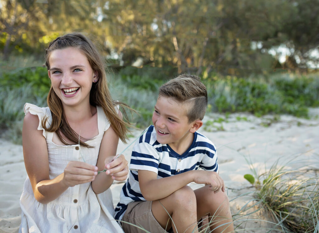 brother and sister sitting on a grassy sand dune laughing