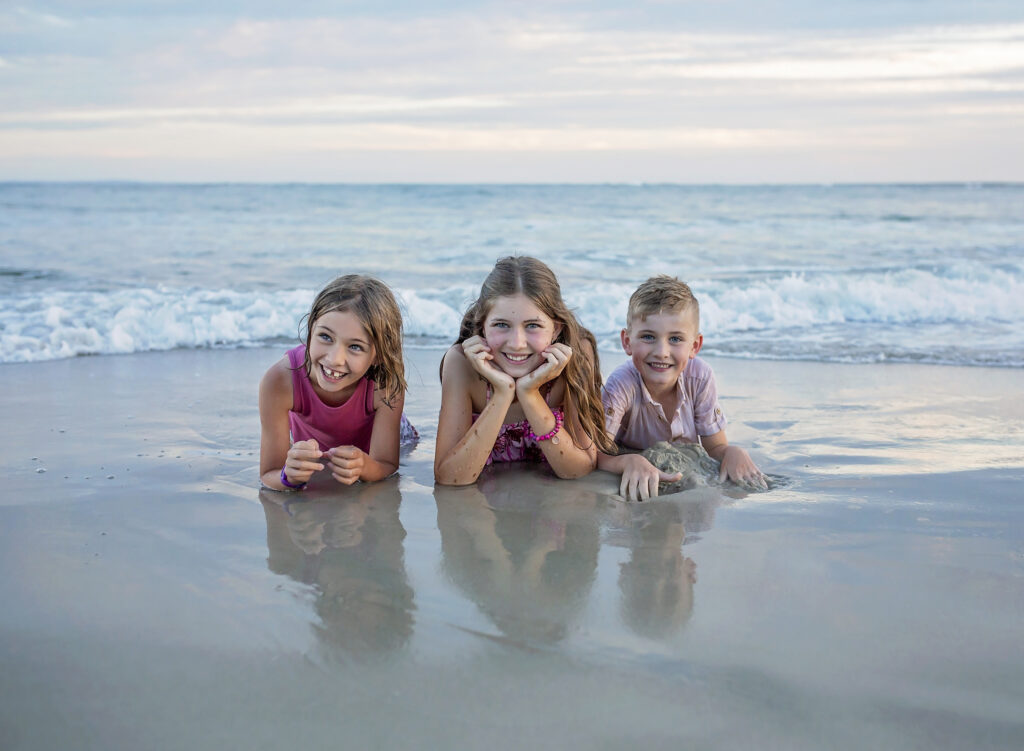 three siblings laying on the beach with the water behind them